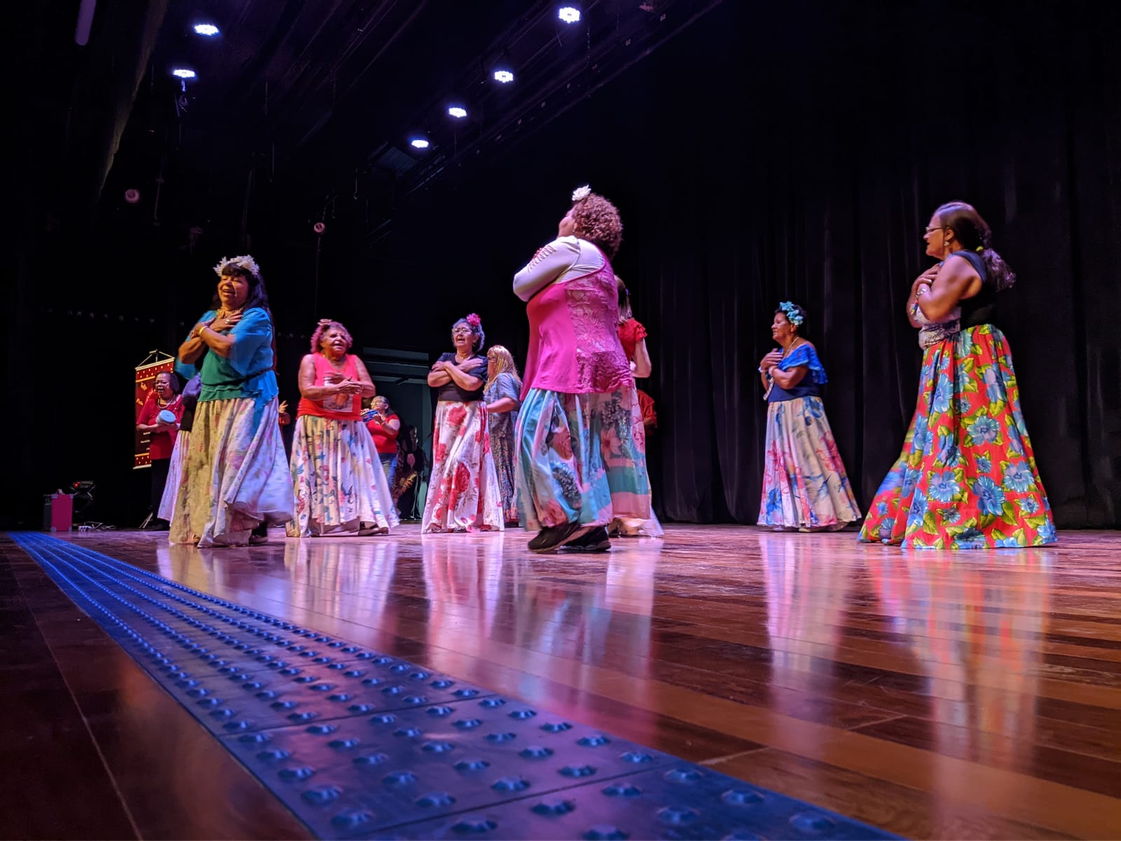 Na imagem, idosas performam dança em cima do palco, elas vestem saias floridas e fazem o gesto de se abraçar.